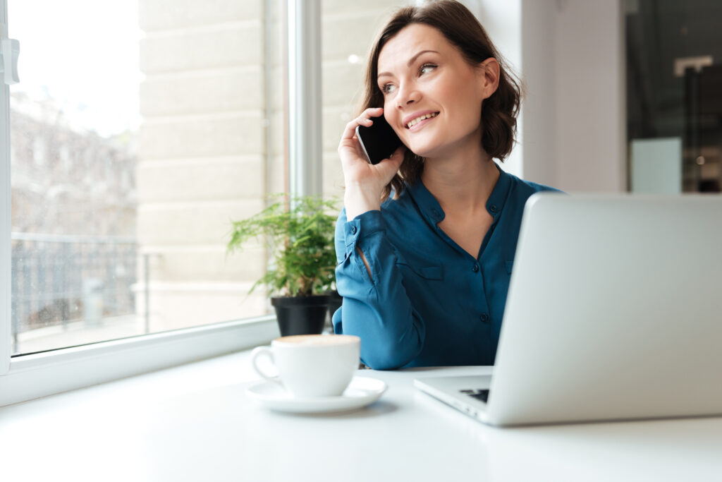 Happy smiling woman talking on mobile phone while sitting at the cafe table with laptop and coffee cup