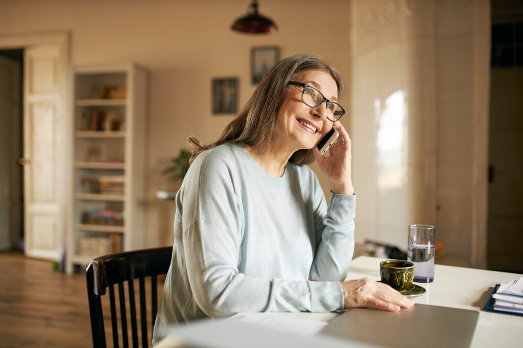 Positive cheerful mature woman pensioner in rectangular eyeglasses using laptop pc sitting at table in cozy home interior, smiling, speaking to friend on smart phone, discussing latest news